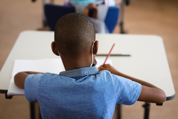 Vista trasera del niño afroamericano estudiando mientras está sentado en su escritorio en la clase en la escuela