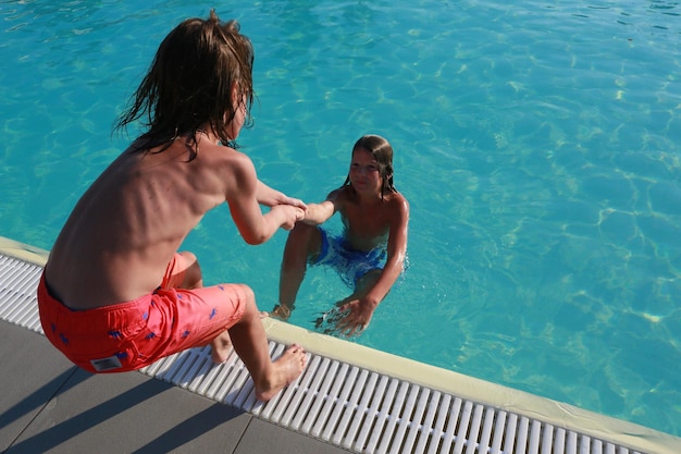 Foto vista trasera de un niño sin camisa sacando a un amigo de la piscina