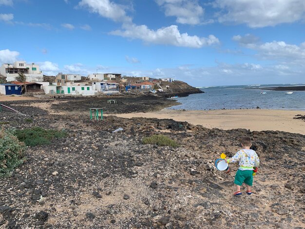 Foto vista trasera de un niño con juguetes caminando por la playa contra el cielo