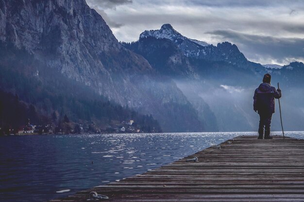 Vista trasera de un niño de pie en el muelle sobre el lago contra la montaña durante el invierno