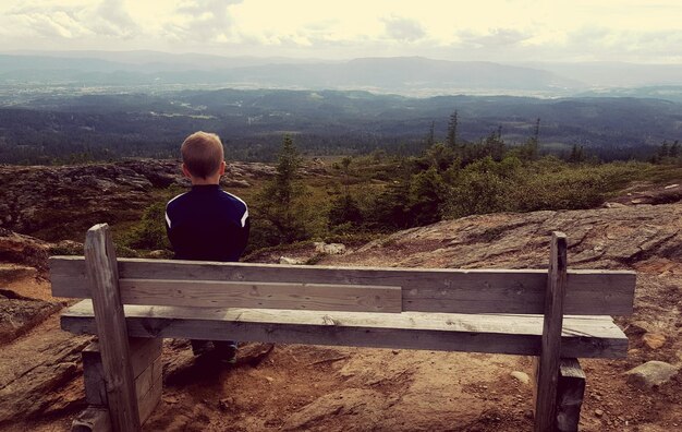 Vista trasera de un niño sentado en un banco en la montaña contra el cielo