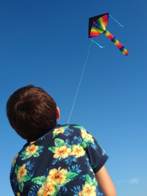 Vista trasera de un niño volando cometa mientras está de pie contra el cielo azul