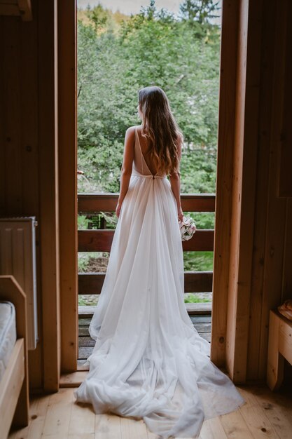 Vista trasera de una novia de cabello castaño vestida con un largo vestido blanco admirando el paisaje montañoso desde...