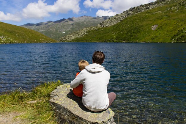 Foto vista trasera de padre e hijo sentados junto al lago contra las montañas