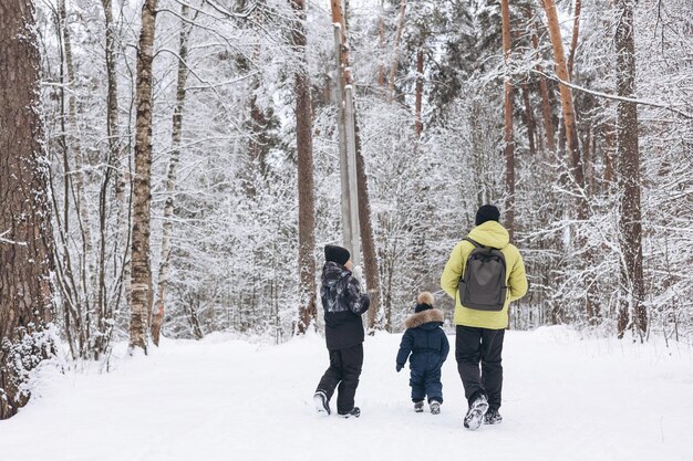 Vista trasera del padre con mochila e hijos pequeños cogidos de la mano caminando juntos en el bosque nevado de invierno. Actividad de invierno al aire libre. Concepto de viaje local y fin de semana familiar.