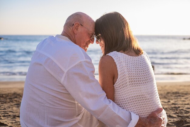 Vista trasera de una pareja de adultos mayores que se abrazan sentados en la playa frente a la puesta de sol disfrutando de sus vacaciones