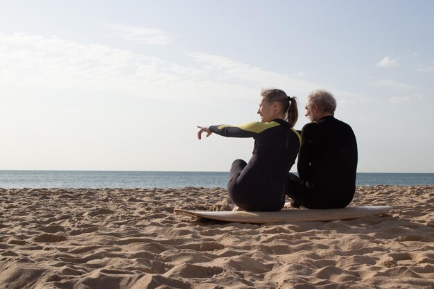 Vista trasera de una pareja de ancianos sentada en una tabla de surf. Hombre y mujer deportivos en trajes de neopreno descansando en una tabla grande en la playa de arena, observando las áreas marinas para surfear. Deporte, estilo de vida saludable para el concepto de personas mayores