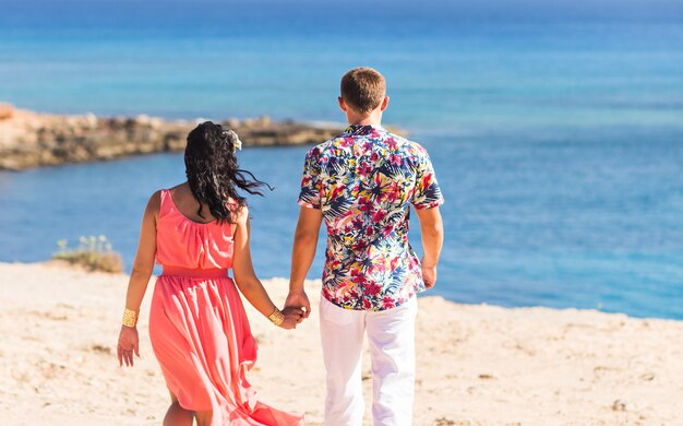 Vista trasera de una pareja caminando por la playa, cogidos de la mano.