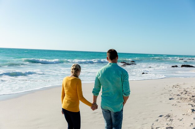 Vista trasera de una pareja caucásica caminando por la playa con el cielo azul y el mar en el fondo, tomados de la mano