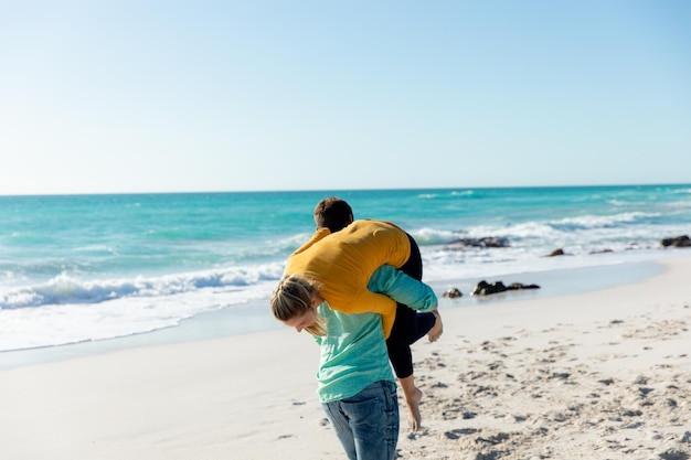 Vista trasera de una pareja caucásica de pie en la playa con el cielo azul y el mar en el fondo, a caballo y divirtiéndose