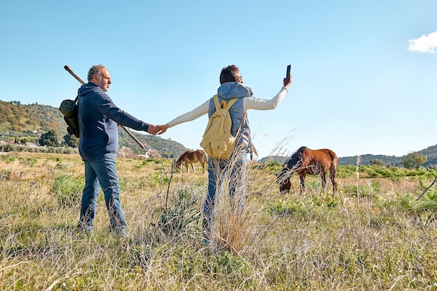 Vista trasera de una pareja cogida de la mano mientras camina por un prado rural con caballos pastando en pastos de otoño o primavera Mujer toma una foto con un teléfono inteligente Unidad con la naturaleza Camino a la montaña