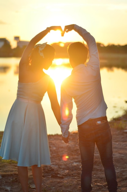 Foto vista trasera de una pareja haciendo forma de corazón mientras están de pie en la orilla del lago al atardecer