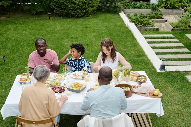 Vista trasera de una pareja madura sentada junto a una mesa servida frente a un joven y feliz hombre y una mujer intercultural y un chico lindo charlando y comiendo