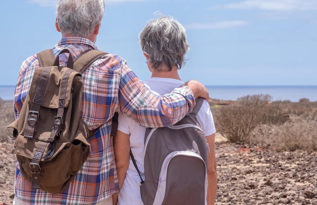 Vista trasera de una pareja senior de viajeros con mochila, disfrutando de una excursión al aire libre. Horizonte sobre el mar y día soleado