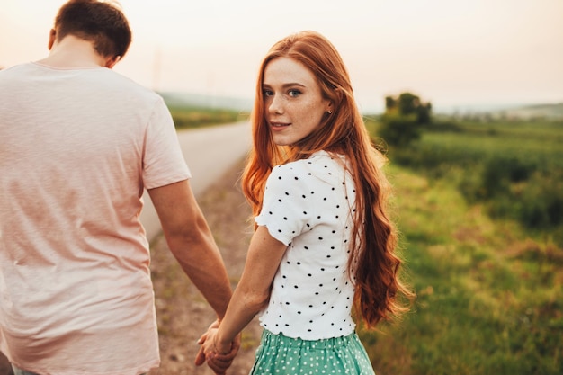 Vista trasera de una pareja tomados de la mano y caminando por un camino rural chica con pelo rojo y pecas tu...