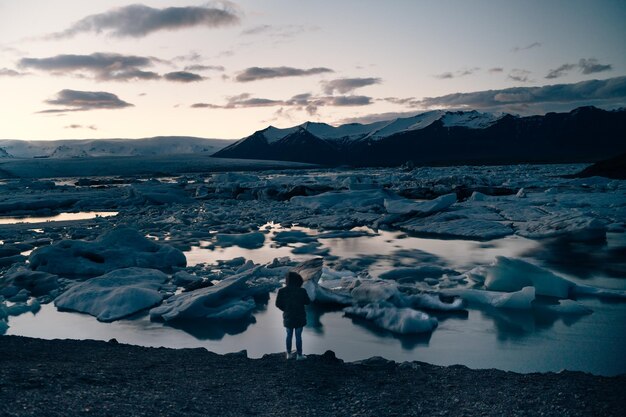 Foto vista trasera de una persona de pie junto al glaciar en el lago contra el cielo durante la puesta de sol