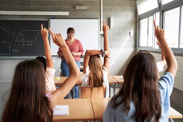 Vista trasera de preadolescentes irreconocibles en el aula levantando las manos para responder a su maestra