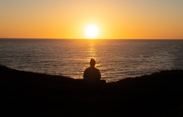 Vista trasera de la silueta de la mujer sentada en el borde de una colina mirando el amanecer