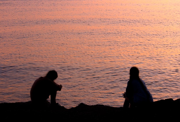 Vista trasera de las siluetas de la pareja sentadas en tierra contra el mar durante la puesta o el amanecer