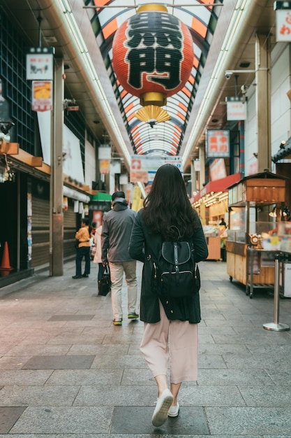 vista trasera con tiro completo mujer asiática caminando bajo una gran linterna roja colgando sobre la arcada en el mercado kuromon ichiba en osaka, japón. traducción:â kuromonâ