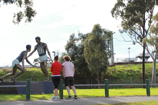 Foto vista trasera a toda longitud de la gente observando la estatua del atleta en el parque contra un cielo despejado