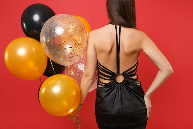 Vista trasera trasera de una mujer joven en un pequeño vestido negro celebrando, sosteniendo globos de aire aislados sobre fondo rojo brillante. Concepto de fiesta de vacaciones de maqueta de cumpleaños de feliz año nuevo de día internacional de la mujer.
