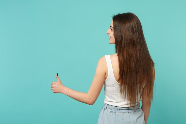 Vista trasera trasera de una mujer joven en ropa casual ligera mirando a un lado, mostrando el pulgar hacia arriba aislado sobre fondo azul turquesa en estudio. Personas sinceras emociones, concepto de estilo de vida. Simulacros de espacio de copia.