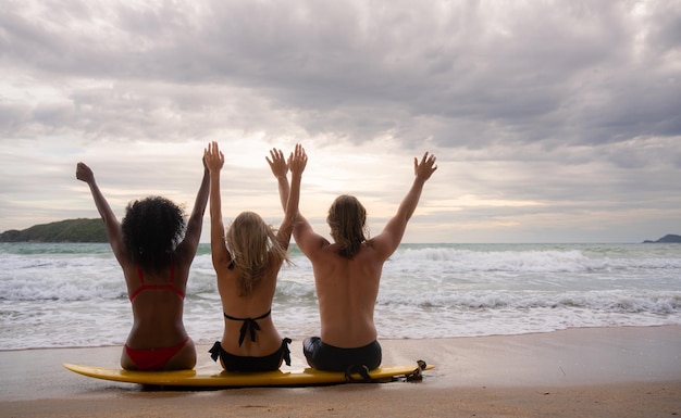 Foto vista trasera de tres amigos sentados en una tabla de surf y mirando al mar