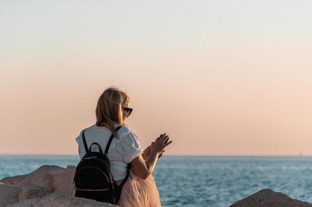 Foto vista trasera de una turista sentada en la costa rocosa tomando fotos de la puesta de sol