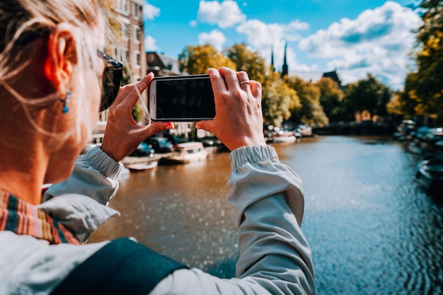 Vista trasera de una turista tomando fotos del canal en Amsterdam en el teléfono móvil en otoño soleado