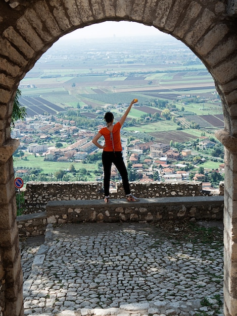 Una vista a través del arco de una niña que está de espaldas en el centro de la foto y mira desde arriba a los techos de casas y campos, su mano está levantada, una naranja está en su mano.