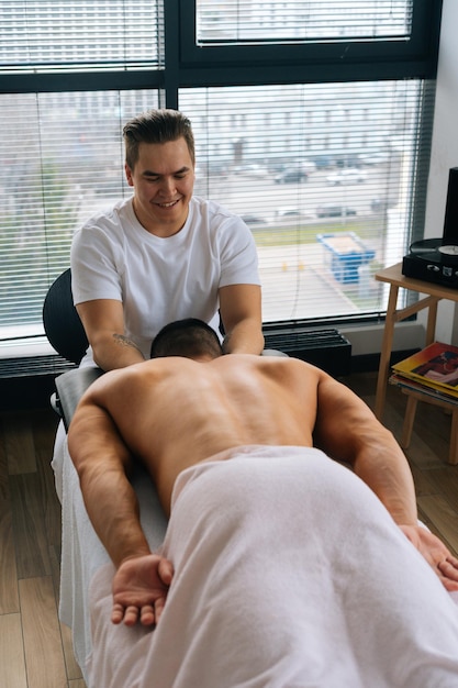 Vista vertical de ángulo alto de masajista masculino profesional con manos fuertes masajeando el hombro de un deportista musculoso acostado boca abajo en la mesa de masaje en un salón de lujo Concepto de estilo de vida saludable