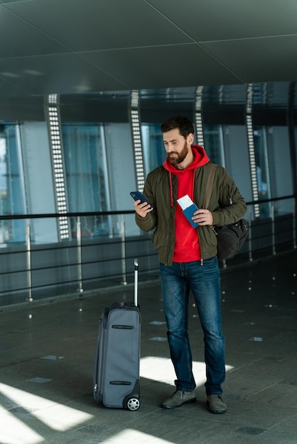 Vista vertical del atractivo hombre sin afeitar parado en el salón del aeropuerto con teléfono y sonriendo mientras revisa algo en la aplicación
