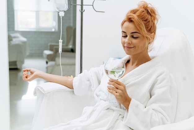 Foto vista vertical de la cintura para arriba de la encantadora mujer en bata de baño blanca sentada en un sillón y recibiendo infusión intravenosa. ella está sosteniendo un vaso de bebida de limón y sonriendo