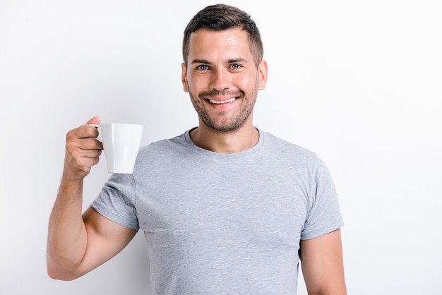 Vista vertical de la cintura para arriba del hombre guapo de pie y sosteniendo una taza de té o café por la mañana y sonriendo mientras mira a la cámara. Foto de stock