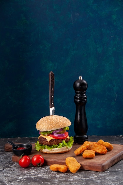 Vista vertical de cuchillo en sabroso sándwich de carne y nuggets de pollo tomates con tallo en tablero de madera salsa ketchup sobre superficie azul oscuro