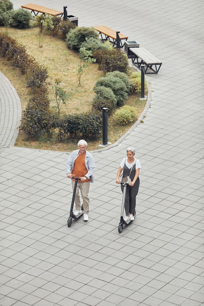 Vista vertical de alto ângulo para casal maduro contemporâneo andando de scooters elétricas no parque em um fundo de azulejos, copie o espaço
