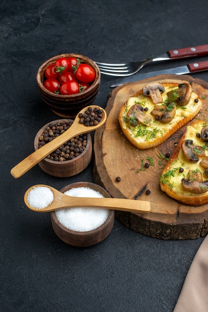 Vista vertical de deliciosos bocadillos con champiñones sobre tablero de madera y cubiertos, tomates pimiento sobre fondo negro