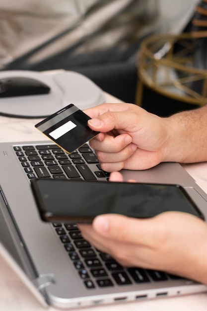 Foto vista vertical del hombre que compra con una tarjeta de crédito negra y su teléfono frente a la computadora portátil, paga y compra en línea