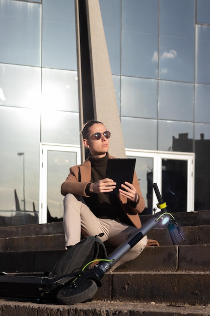 Vista vertical de un joven y apuesto hombre de negocios leyendo un libro electrónico en su tableta en la escalera con un scooter eléctrico