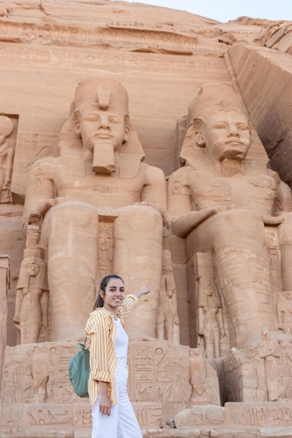 Vista vertical de una joven turista señalando el templo de Abu Simbel desde afuera con traje de turista y mirando a la cámara