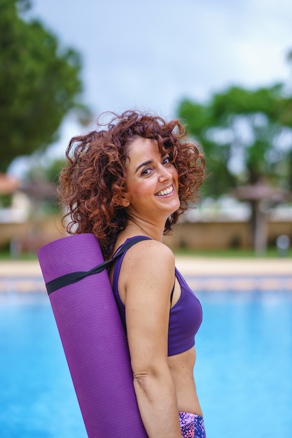 Vista vertical del retrato de mujer de pelo rizado con sonrisa blanca vestida con un traje deportivo sosteniendo una estera de yoga rosa. Estilo de vida fitness, ejercicio y hábitos saludables en verano.