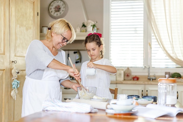 Visualização horizontal. Menina bonita encaracolada com sua avó feliz cozinhando juntos na mesa da cozinha.