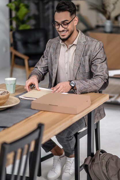 Visualización de correspondencia. Elegante joven barbudo con anteojos sentado a la mesa en casa mirando con placer la correspondencia