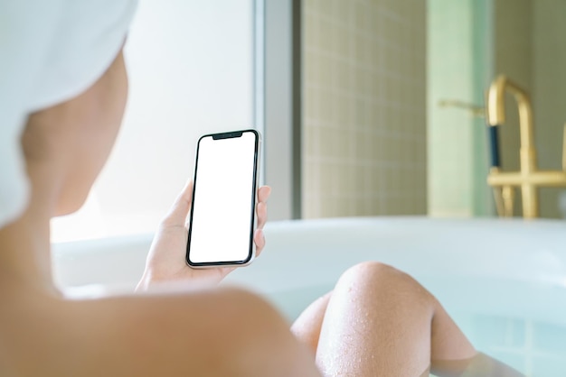 Foto visualización de la pantalla en blanco del teléfono en la mano de la mujer, relajarse en la bañera con el teléfono inteligente en la mañana