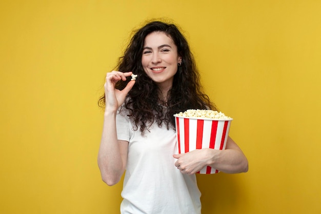 Visualizador de garota encaracolada em camiseta branca segura e come pipoca em fundo amarelo