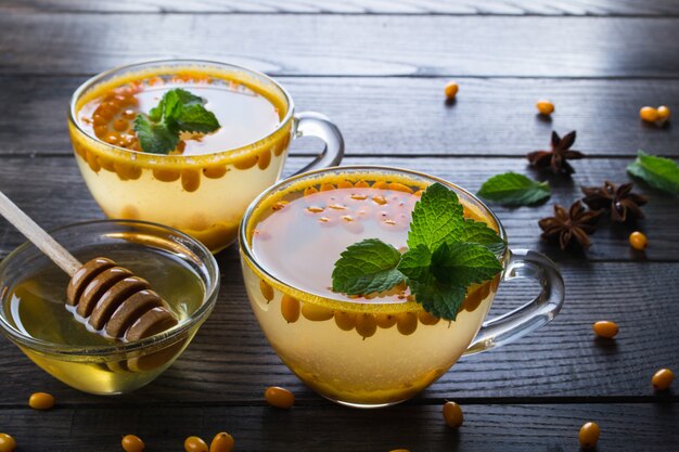 Vitamin gesunder Sanddorn-Tee in Glasbechern mit frischen rohen Sanddornbeeren und Zimtstangen, Anissternen, Minze und Honig auf einem dunklen Küchentisch.