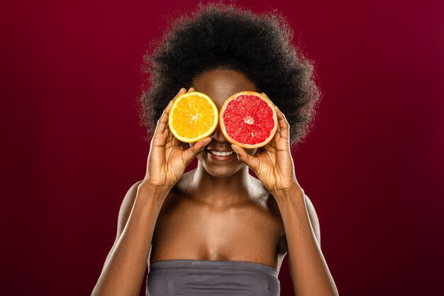 Vitamina C. Bonita mujer alegre sosteniendo dos mitades de frutas mientras está de pie contra la pared roja