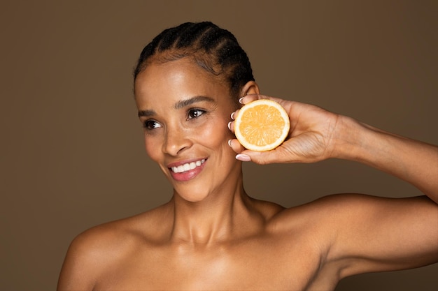 Vitamina C en la rutina de cuidado facial feliz mujer negra de mediana edad sosteniendo la mitad de naranja y sonriendo