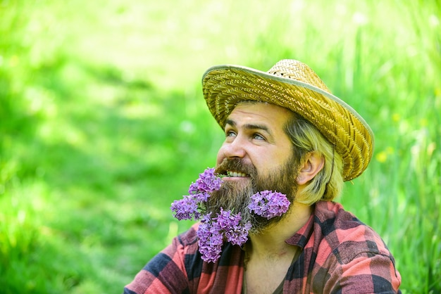 Viva brilhantemente. agricultor relaxado com chapéu de palha. jardineiro homem maduro relaxe na grama verde. aproveite a natureza da primavera. temporada de verão com lilás. Hipster de fazenda brutal tem uma barba longa e saudável. flor no cabelo.